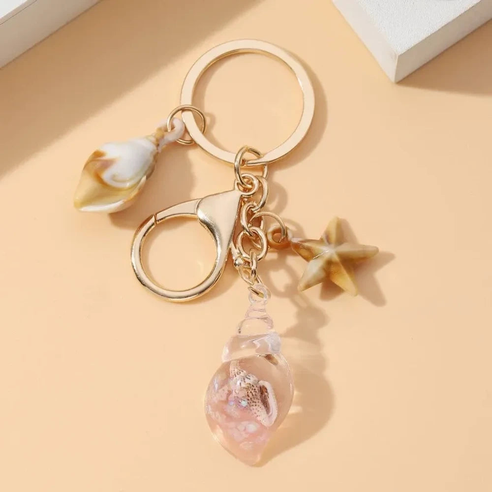 Six ocean-themed keychain charms featuring resin crystal shells in green, purple, blue, clear, and pink, with mini starfish and natural shell accents on gold-tone rings and lobster clasps. - 12