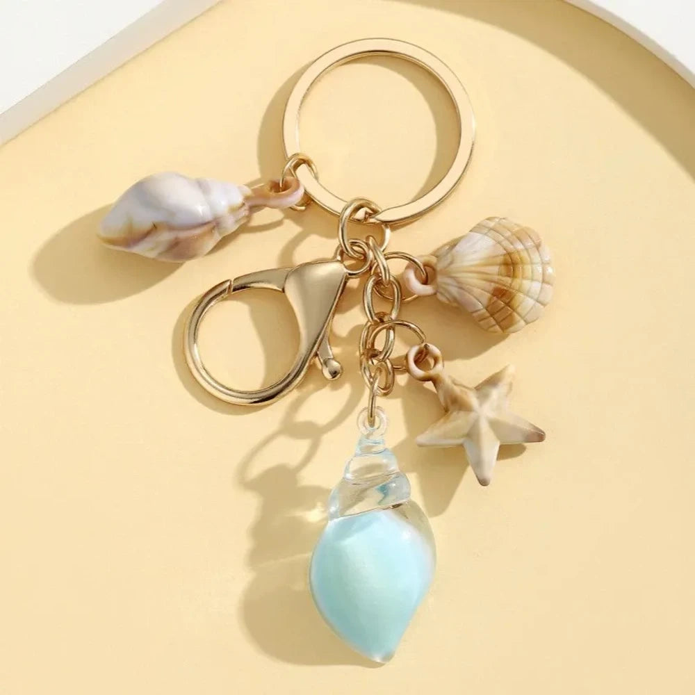 Six ocean-themed keychain charms featuring resin crystal shells in green, purple, blue, clear, and pink, with mini starfish and natural shell accents on gold-tone rings and lobster clasps. - 13