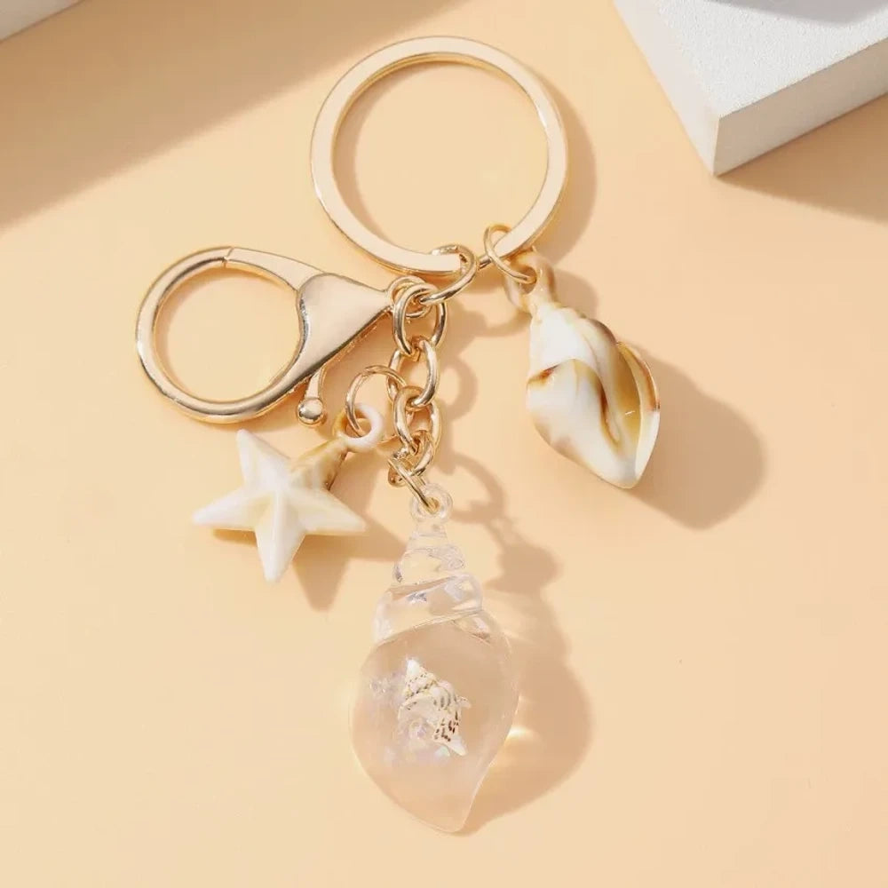 Six ocean-themed keychain charms featuring resin crystal shells in green, purple, blue, clear, and pink, with mini starfish and natural shell accents on gold-tone rings and lobster clasps. - 3