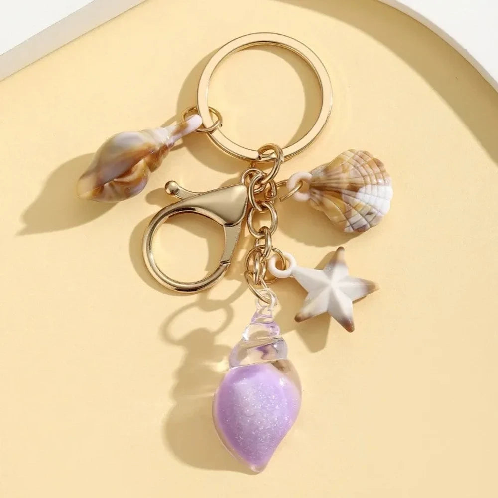 Six ocean-themed keychain charms featuring resin crystal shells in green, purple, blue, clear, and pink, with mini starfish and natural shell accents on gold-tone rings and lobster clasps. - 6