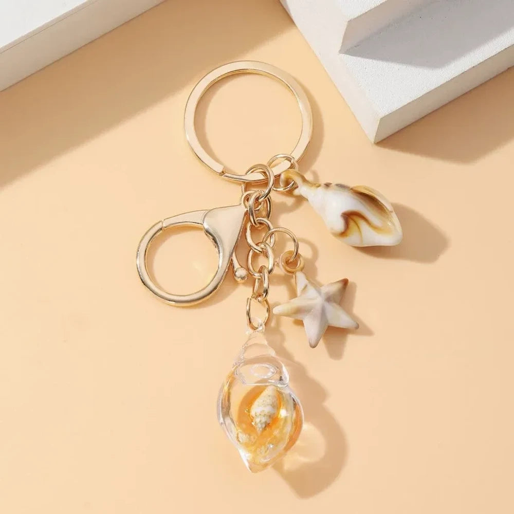 Six ocean-themed keychain charms featuring resin crystal shells in green, purple, blue, clear, and pink, with mini starfish and natural shell accents on gold-tone rings and lobster clasps. - 7