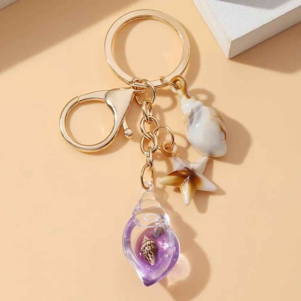 Six ocean-themed keychain charms featuring resin crystal shells in green, purple, blue, clear, and pink, with mini starfish and natural shell accents on gold-tone rings and lobster clasps. - 9