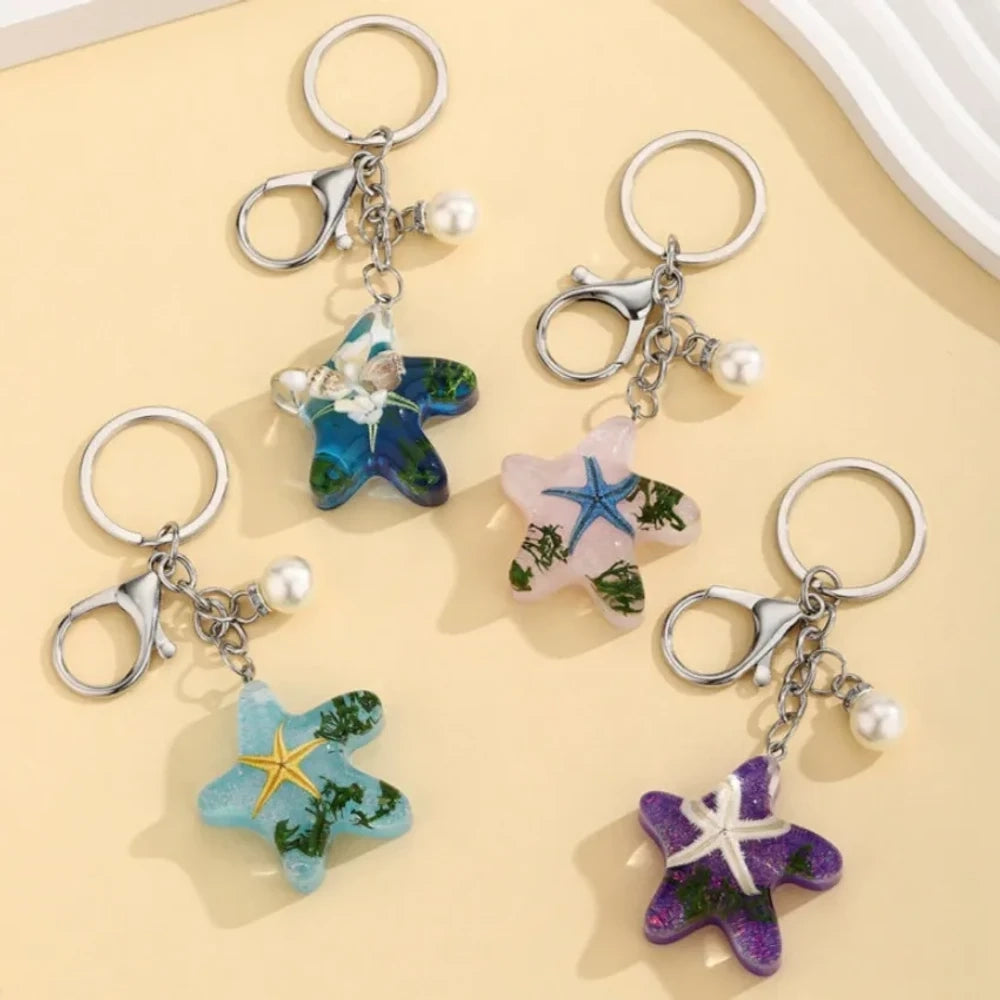 Four ocean starfish-shaped resin keychain charms in blue, pink, light blue, and purple with embedded starfish, flowers, and seaweed, each with a pearl accent and silver lobster clasp ring.