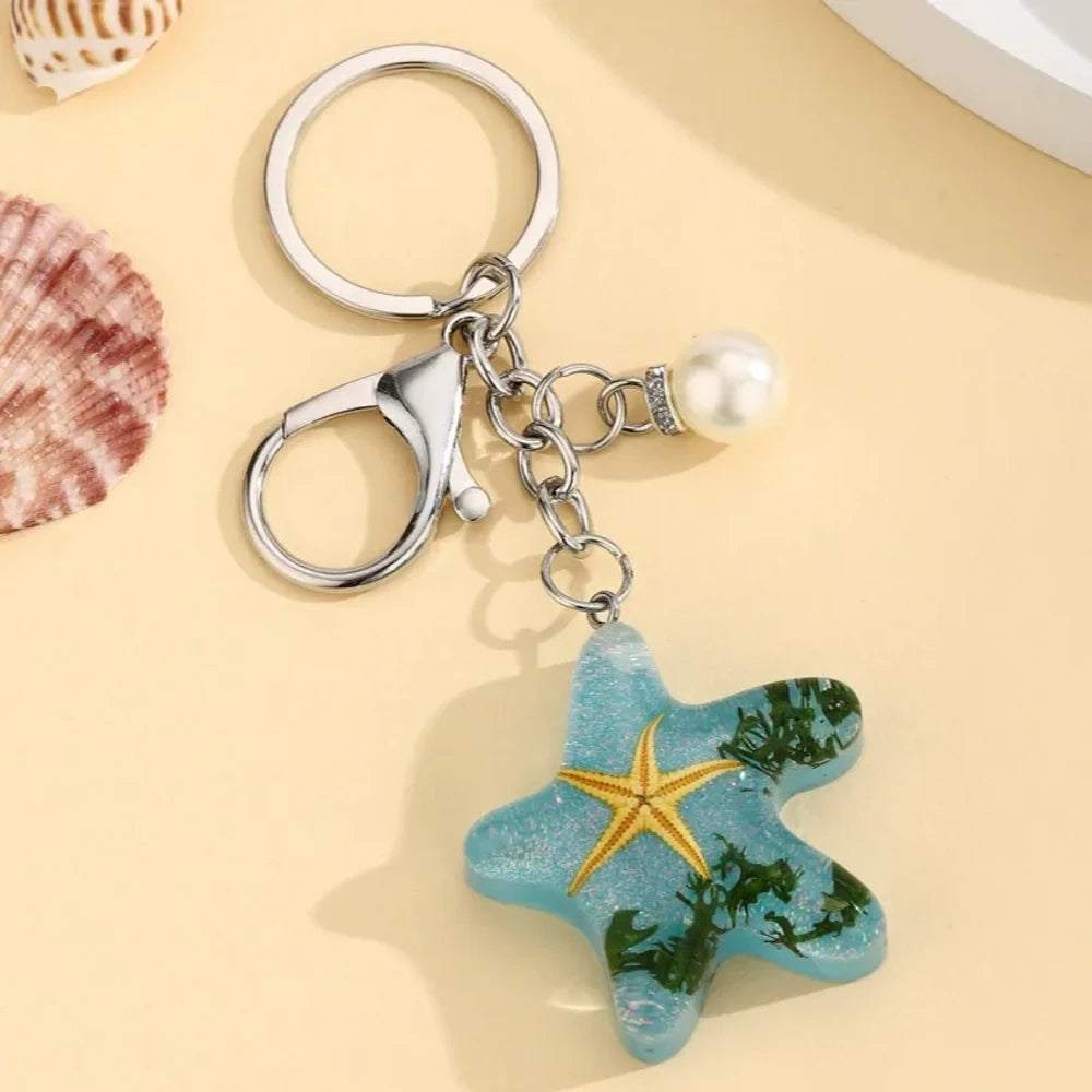 Four ocean starfish-shaped resin keychain charms in blue, pink, light blue, and purple with embedded starfish, flowers, and seaweed, each with a pearl accent and silver lobster clasp ring. - Light Blue
