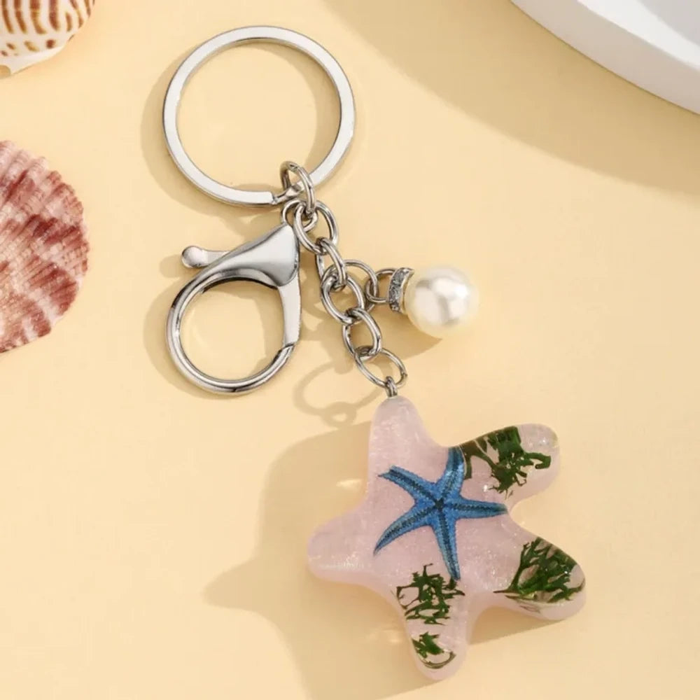 Four ocean starfish-shaped resin keychain charms in blue, pink, light blue, and purple with embedded starfish, flowers, and seaweed, each with a pearl accent and silver lobster clasp ring. - Pink