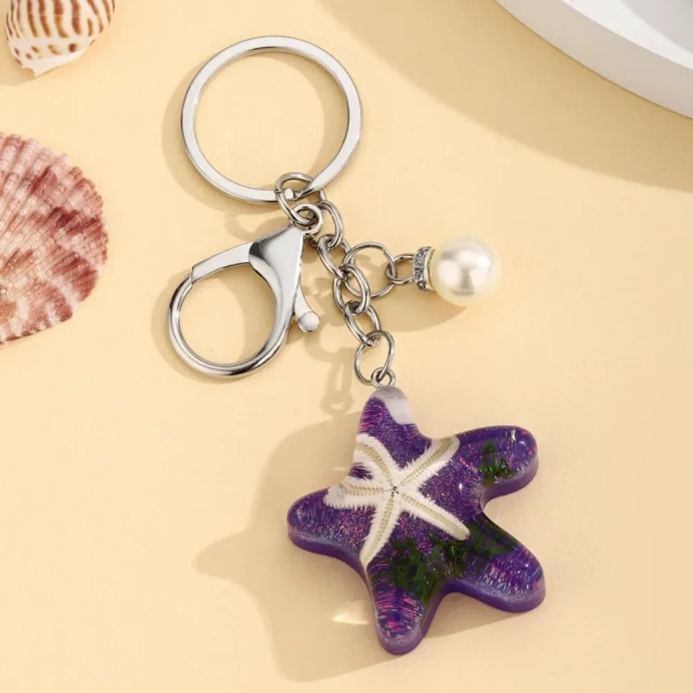 Four ocean starfish-shaped resin keychain charms in blue, pink, light blue, and purple with embedded starfish, flowers, and seaweed, each with a pearl accent and silver lobster clasp ring. - Purple