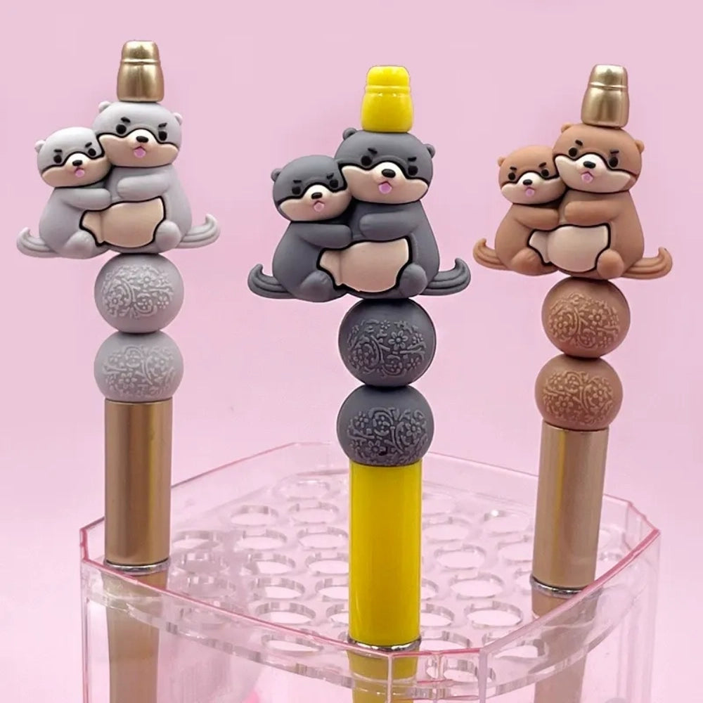 Three 3D silicone focal beads featuring cute otter pairs in gray, dark gray, and brown colorways, each topped on stacked decorative beads and inserted into beadable pens, displayed in a clear acrylic holder against a pink background.