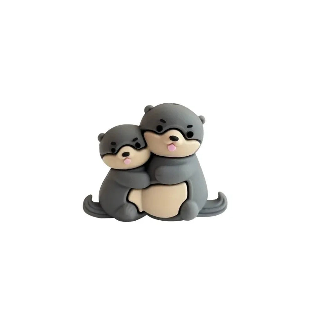 Three 3D silicone focal beads featuring cute otter pairs in gray, dark gray, and brown colorways, each topped on stacked decorative beads and inserted into beadable pens, displayed in a clear acrylic holder against a pink background. - Dark Gray