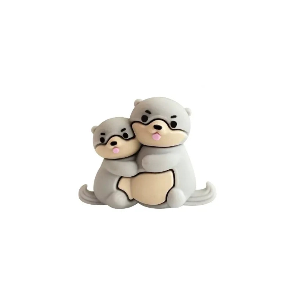 Three 3D silicone focal beads featuring cute otter pairs in gray, dark gray, and brown colorways, each topped on stacked decorative beads and inserted into beadable pens, displayed in a clear acrylic holder against a pink background. - Light Gray