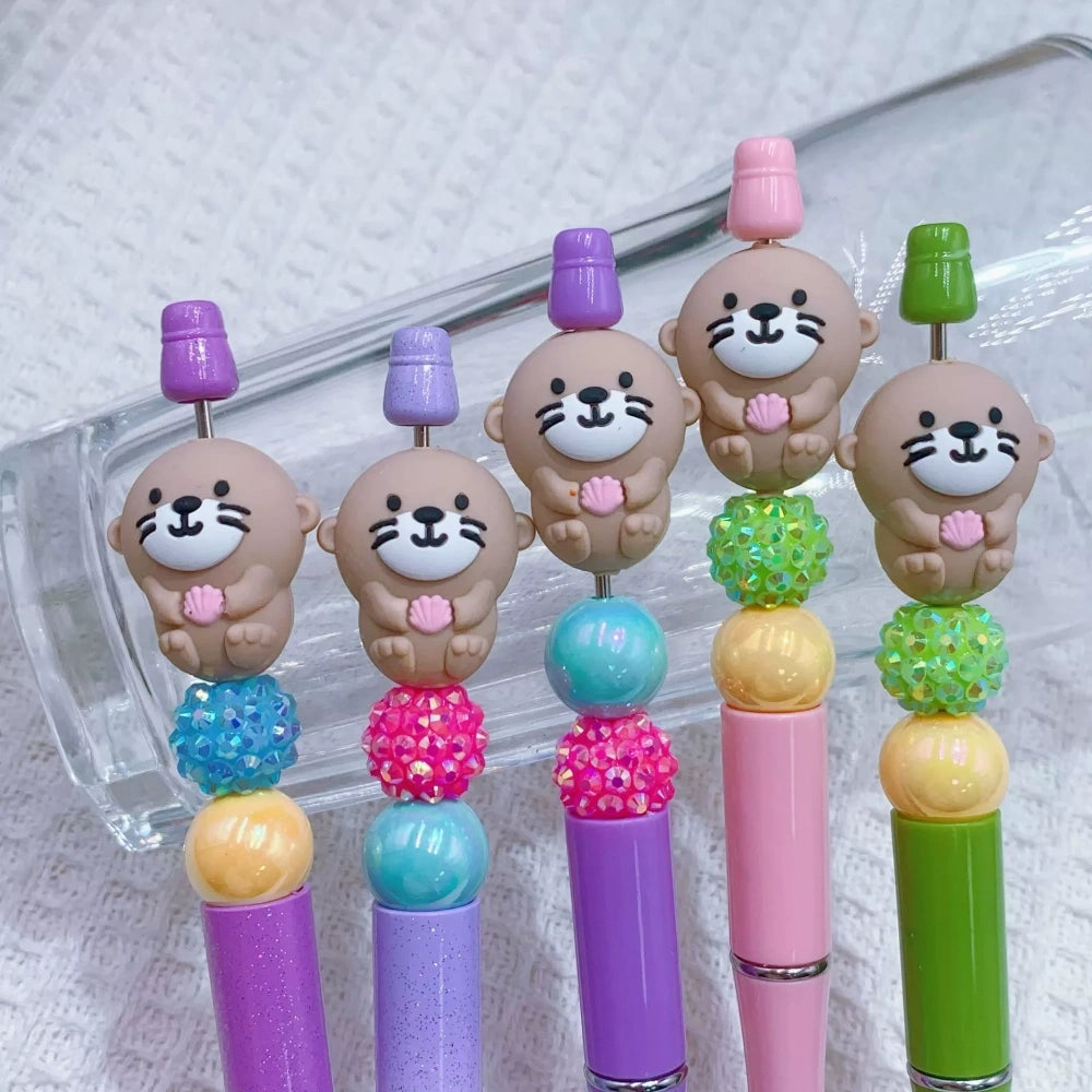Five cute otter silicone focal beads holding shells, displayed on colorful beadable pens in purple, lavender, pink, and green with rhinestone and pearl accent beads.