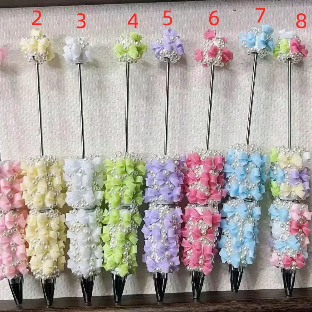 Set of 7 pastel-colored beadable rhinestone pens decorated with satin ribbon bows and pearl beads in yellow, white, green, purple, pink, blue, and multicolor variants displayed upright on a textured background.