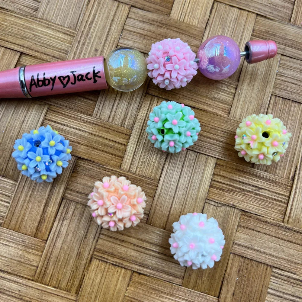 Assorted pastel floral cluster ball resin fancy beads in pink, blue, green, peach, yellow, and white with rhinestone accents, displayed alongside a pink beadable pen on a wicker surface.