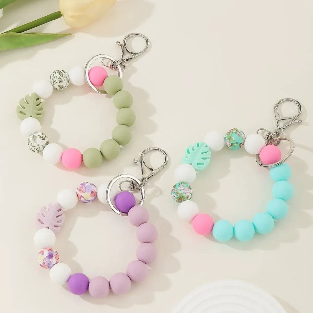 Three pastel silicone bead bracelet keychains in sage green, lavender, and teal colorways featuring floral print beads, monstera leaf silicone beads, pink accent beads, white beads, and silver lobster clasp hardware on a cream background.