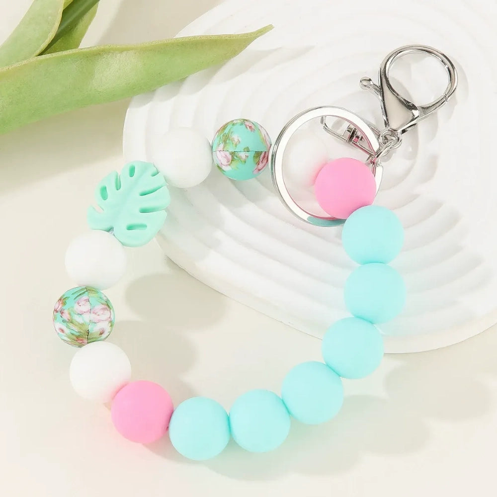 Three pastel silicone bead bracelet keychains in sage green, lavender, and teal colorways featuring floral print beads, monstera leaf silicone beads, pink accent beads, white beads, and silver lobster clasp hardware on a cream background. - Blue