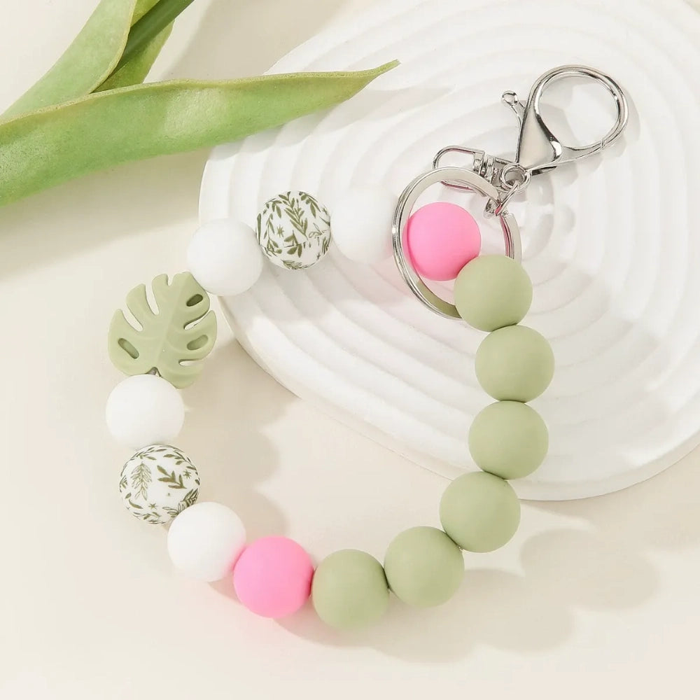Three pastel silicone bead bracelet keychains in sage green, lavender, and teal colorways featuring floral print beads, monstera leaf silicone beads, pink accent beads, white beads, and silver lobster clasp hardware on a cream background. - Green