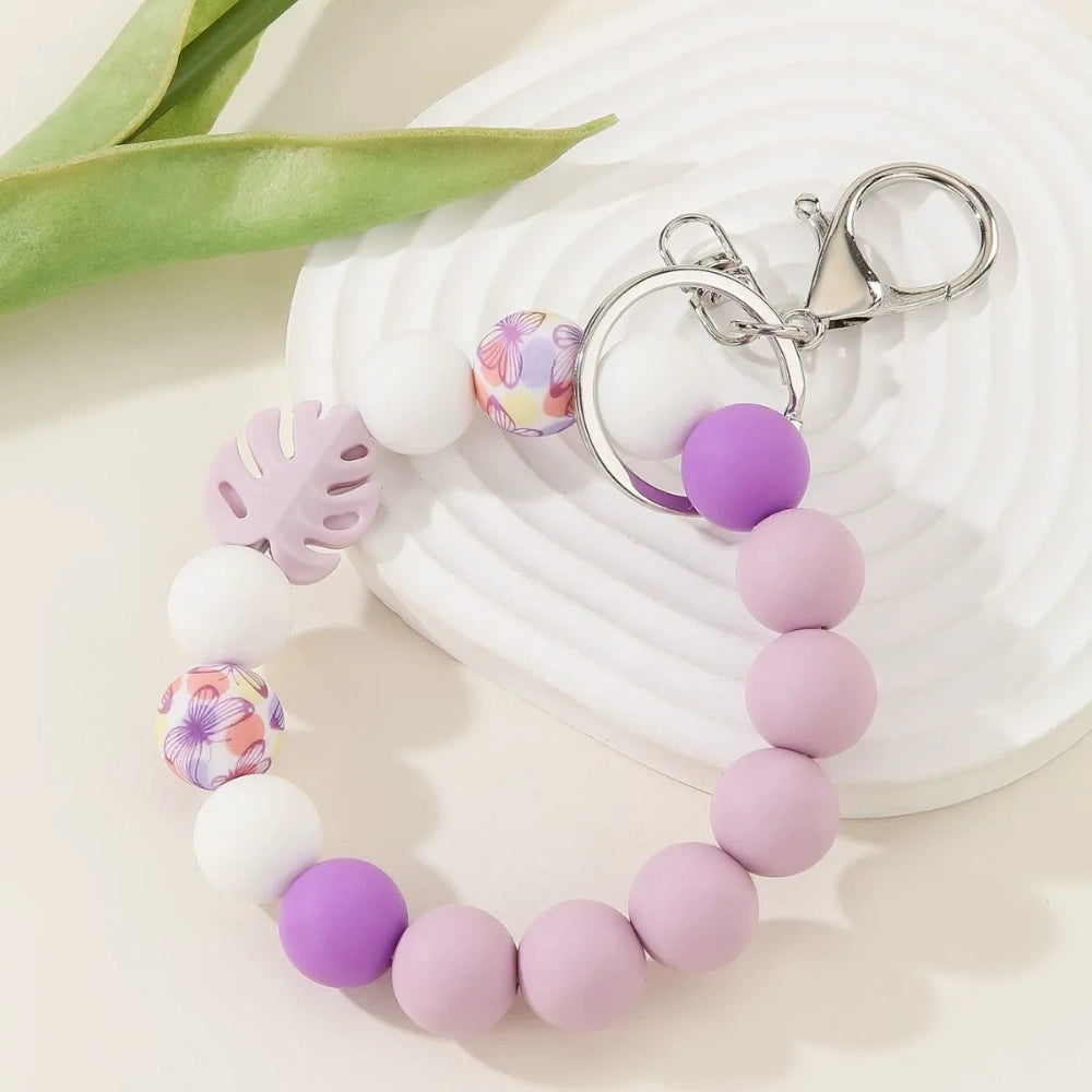 Three pastel silicone bead bracelet keychains in sage green, lavender, and teal colorways featuring floral print beads, monstera leaf silicone beads, pink accent beads, white beads, and silver lobster clasp hardware on a cream background. - Purple