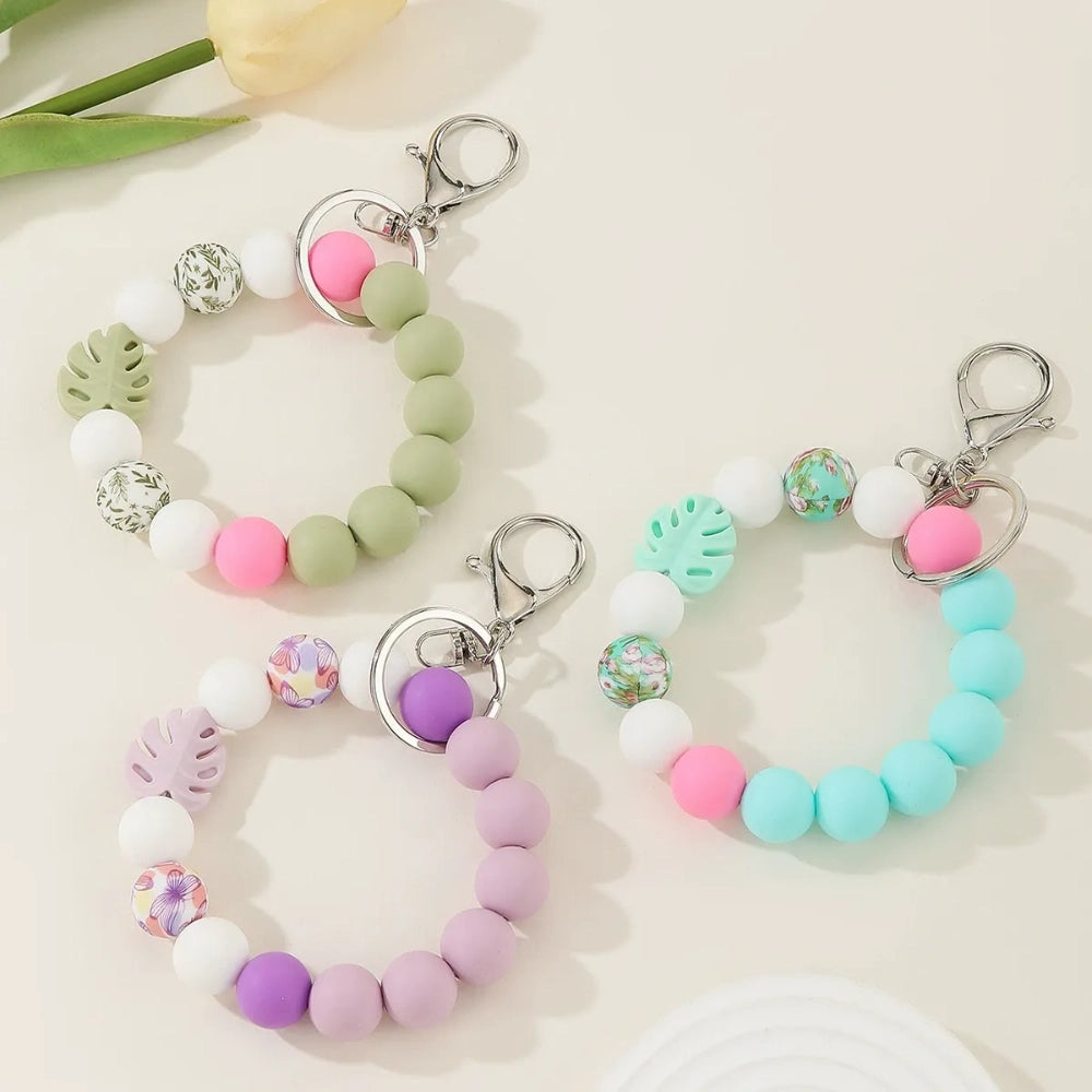 Three pastel silicone bead bracelet keychains in sage green, lavender, and teal colorways featuring floral print beads, monstera leaf silicone beads, pink accent beads, white beads, and silver lobster clasp hardware on a cream background. - Random Mixed