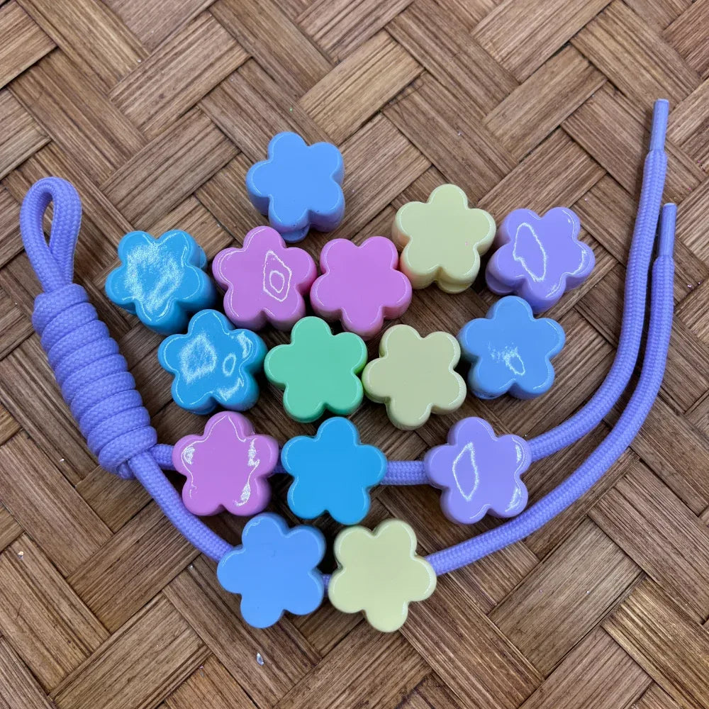 Pastel flower-shaped acrylic beads in blue, purple, pink, mint, and yellow-green colors displayed on a woven surface with a purple cord, ideal for beadable pens, keychains, and DIY jewelry making.