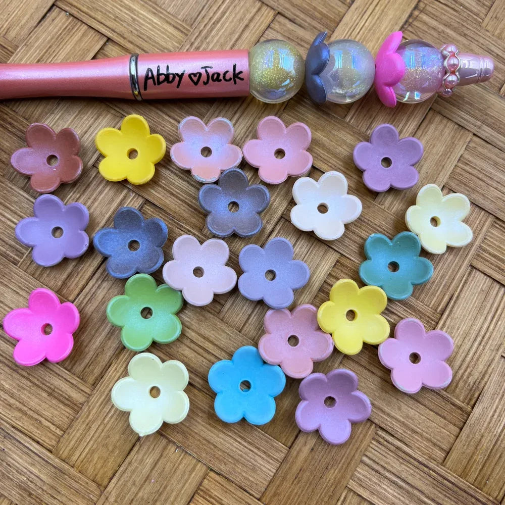 Assorted pastel flower-shaped acrylic spacer beads in pink, purple, blue, yellow, green, and white displayed on a woven surface next to a beadable pen labeled Abby Jack.