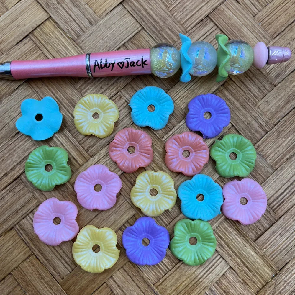 Assorted pastel flower-shaped acrylic spacer beads in blue, pink, yellow, green, and purple displayed next to a beadable pen on a wicker surface.