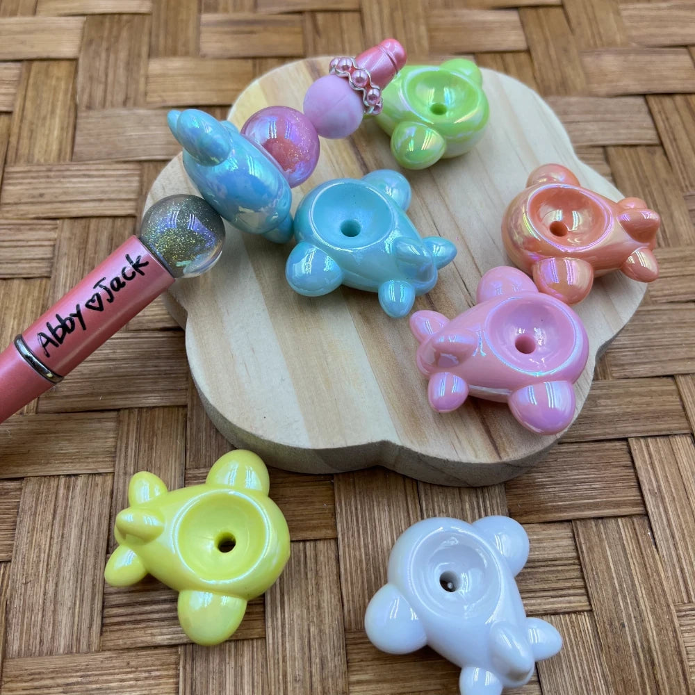 Assorted pastel iridescent ceramic bear flower bead caps in pink, blue, green, orange, yellow, and white displayed on a wooden tray with a beadable pen, for DIY jewelry making and crafts.