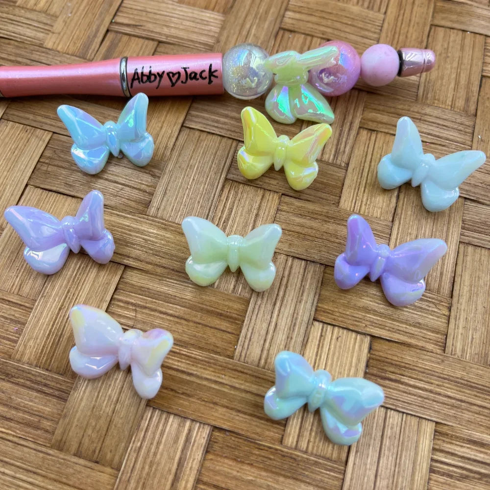 Assorted pastel iridescent butterfly-shaped acrylic beads in purple, yellow, mint, blue, and pink displayed on a woven surface next to a beadable pen.