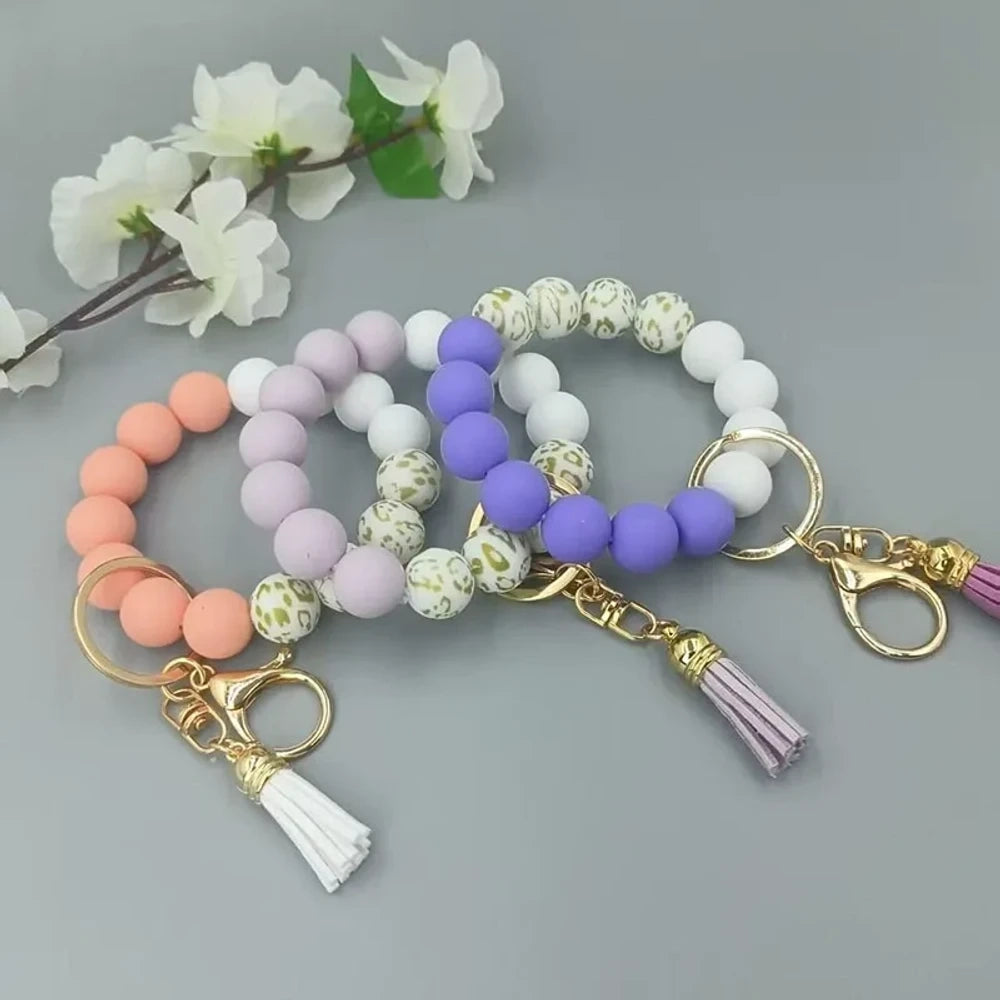 Pastel silicone bead bracelets in peach, lavender, and purple with leopard print beads, gold lobster clasps, and tassel charms on a gray background.