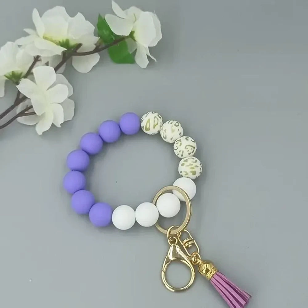 Pastel silicone bead bracelets in peach, lavender, and purple with leopard print beads, gold lobster clasps, and tassel charms on a gray background. - Dark Purple