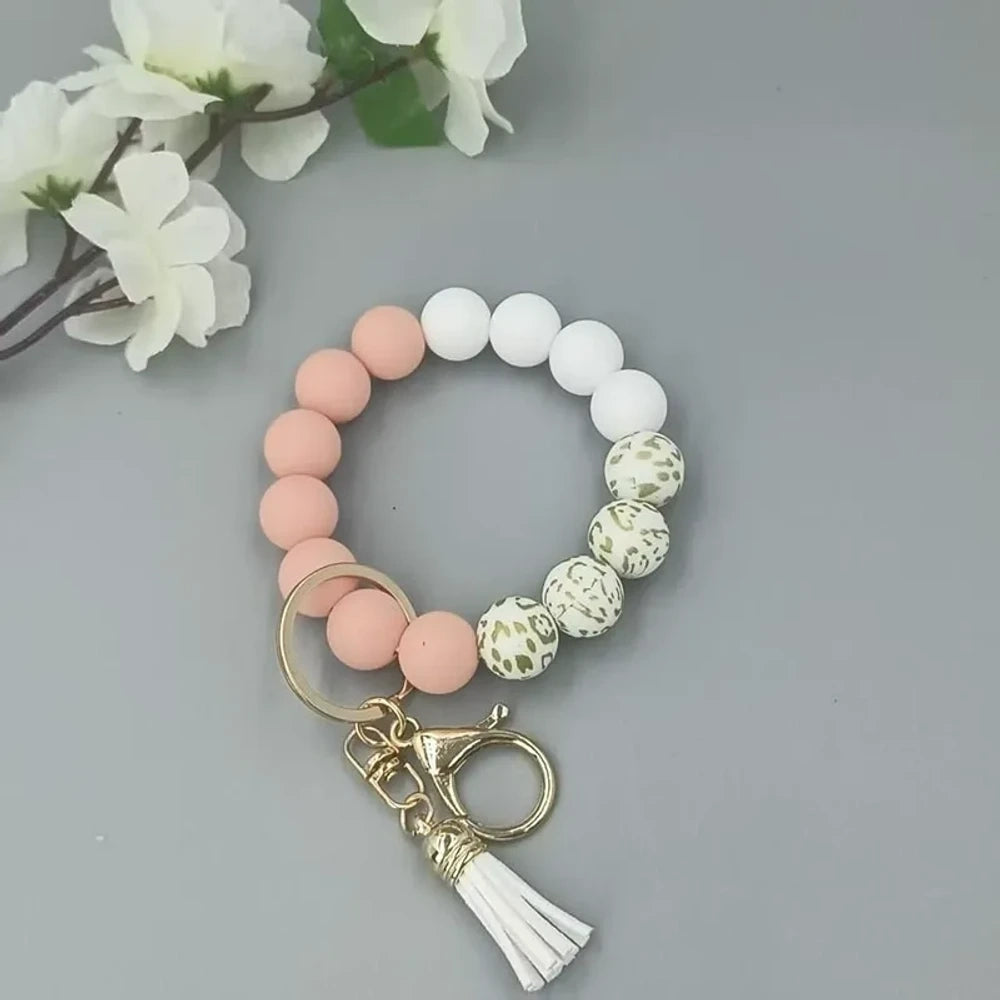 Pastel silicone bead bracelets in peach, lavender, and purple with leopard print beads, gold lobster clasps, and tassel charms on a gray background. - Orange