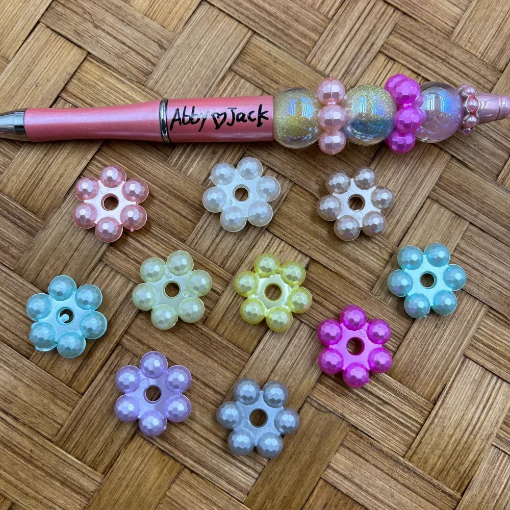 Assorted pastel pearl flower-shaped acrylic spacer beads in pink, white, blue, yellow, purple, gray and hot pink, displayed on a woven surface next to a beaded pen.