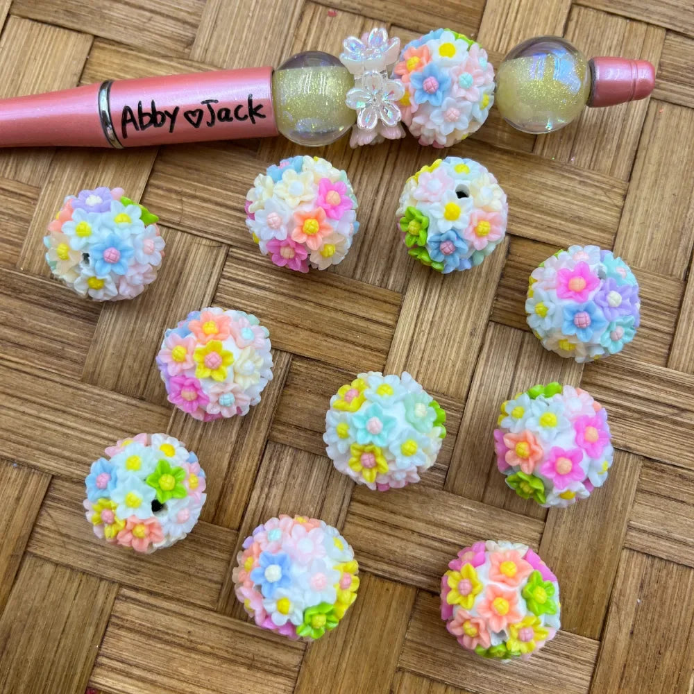 Pastel rainbow flower cluster resin beads in pink, blue, yellow, and green displayed on a wicker surface next to a decorated beadable pen, perfect for DIY jewelry making and beadable pens.