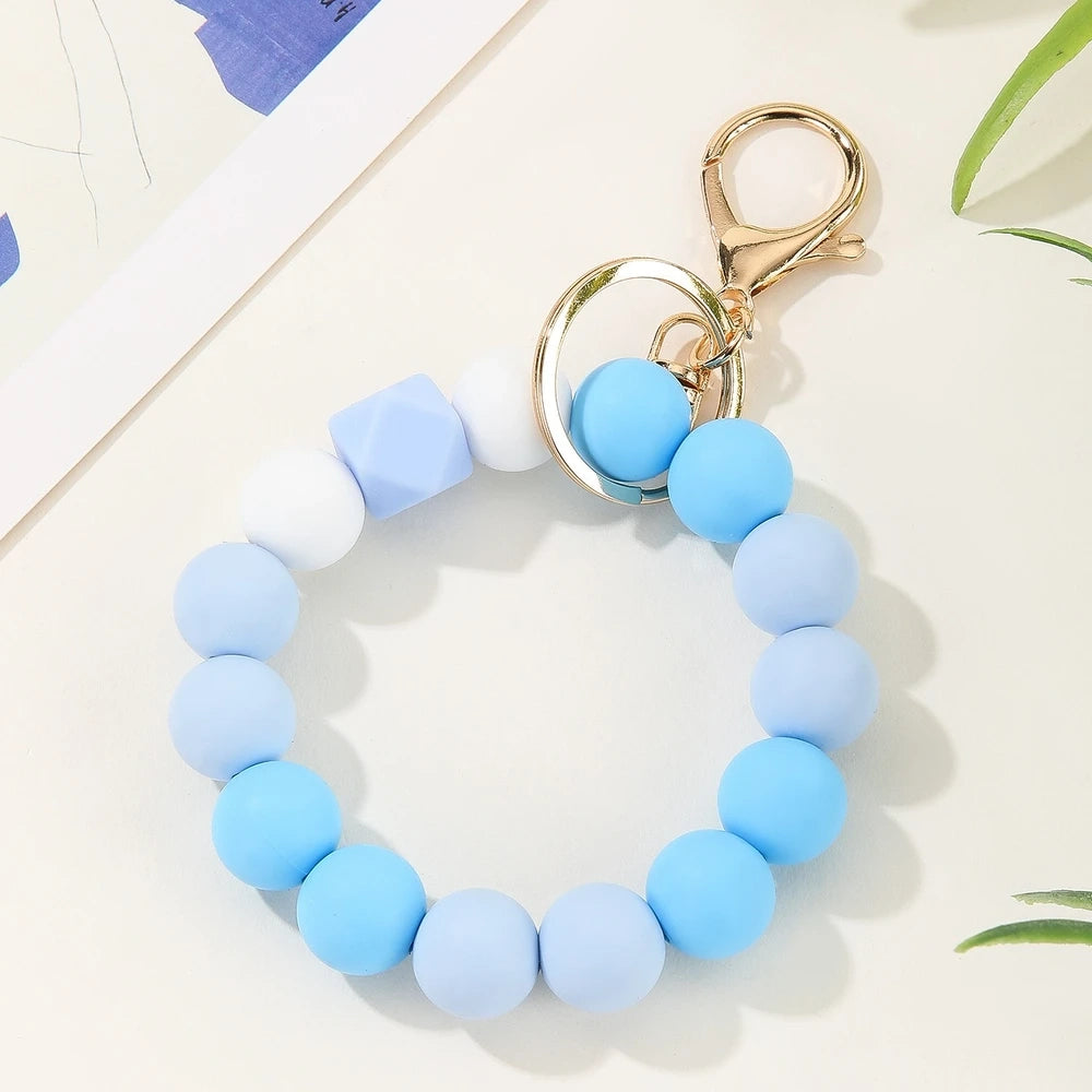 Four pastel rainbow silicone bead keychain bracelets in orange-yellow, mint-yellow, pink-purple, and blue colorways with gold lobster clasp hardware on a white marble surface. - Blue