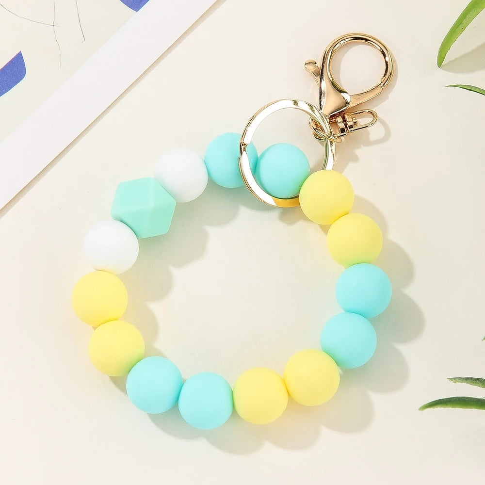 Four pastel rainbow silicone bead keychain bracelets in orange-yellow, mint-yellow, pink-purple, and blue colorways with gold lobster clasp hardware on a white marble surface. - Green