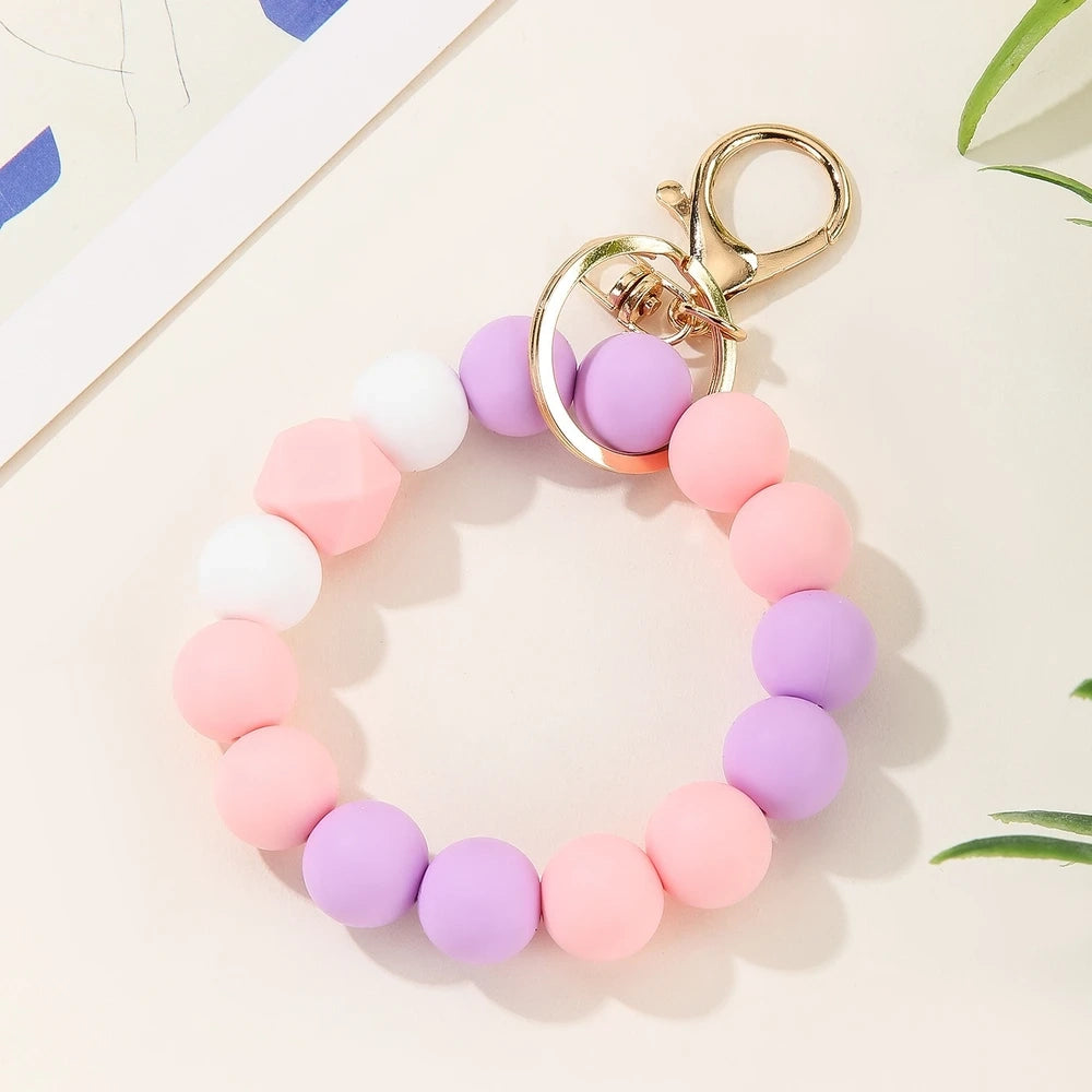 Four pastel rainbow silicone bead keychain bracelets in orange-yellow, mint-yellow, pink-purple, and blue colorways with gold lobster clasp hardware on a white marble surface. - Purple