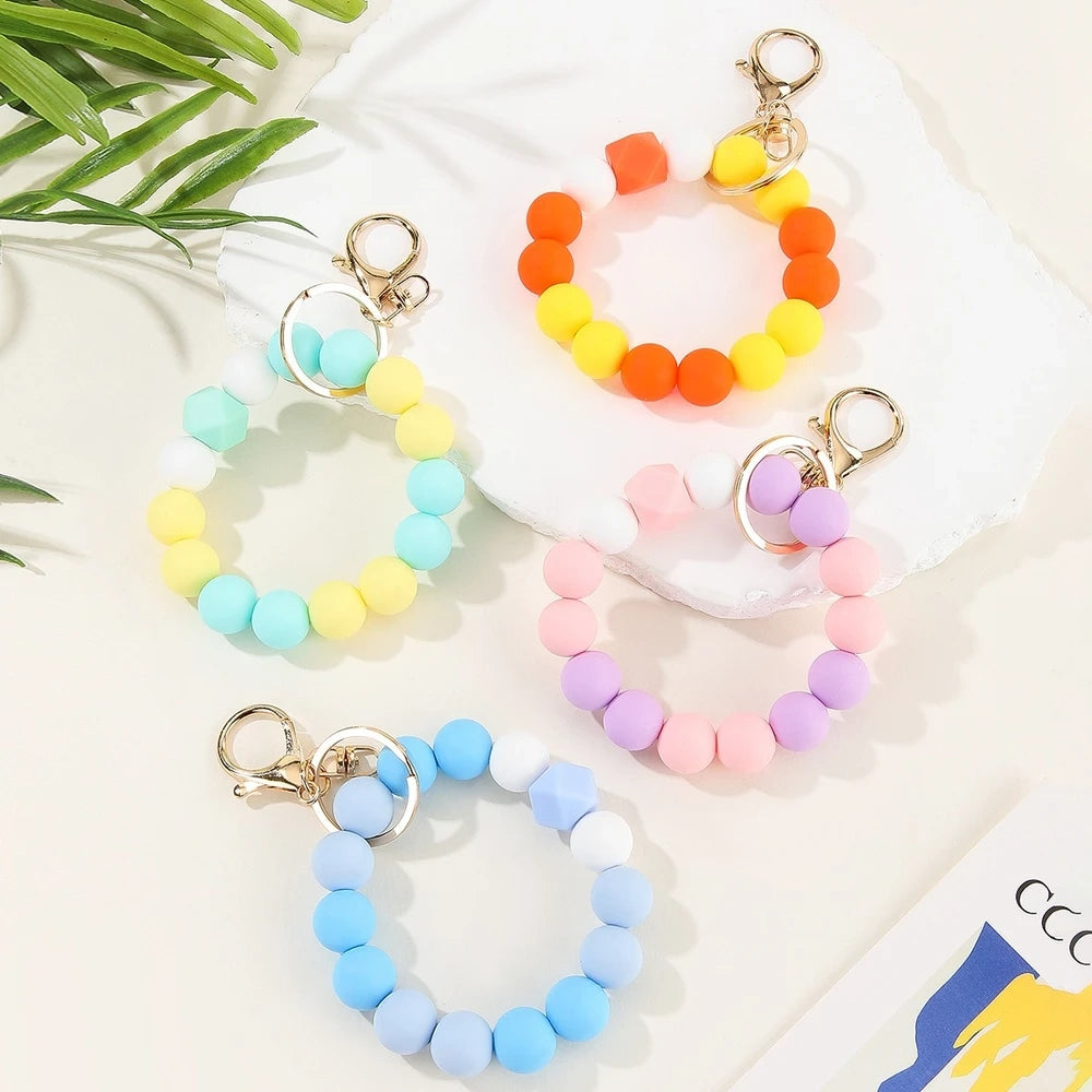 Four pastel rainbow silicone bead keychain bracelets in orange-yellow, mint-yellow, pink-purple, and blue colorways with gold lobster clasp hardware on a white marble surface. - Random Mixed