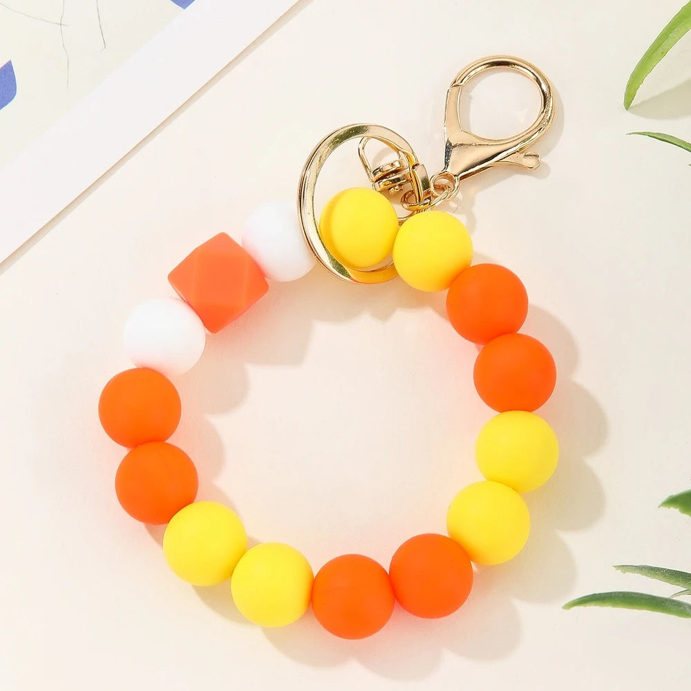 Four pastel rainbow silicone bead keychain bracelets in orange-yellow, mint-yellow, pink-purple, and blue colorways with gold lobster clasp hardware on a white marble surface. - Red