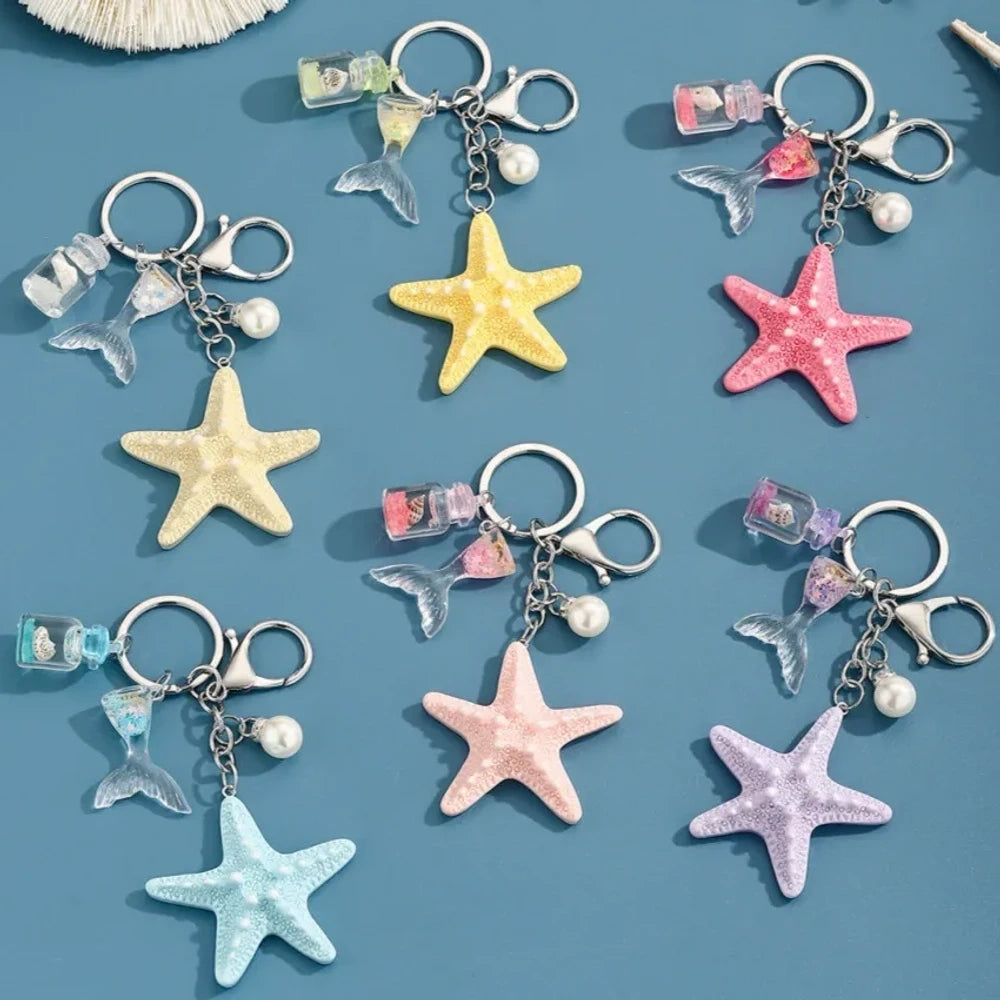 Six pastel resin starfish keychain charms in white, yellow, pink, blue, light pink, and purple, each featuring a mermaid tail, pearl bead, and mini bottle accent on silver chain rings, displayed on a teal background.