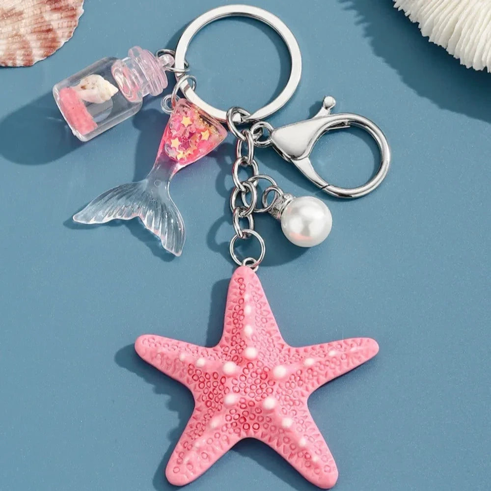 Six pastel resin starfish keychain charms in white, yellow, pink, blue, light pink, and purple, each featuring a mermaid tail, pearl bead, and mini bottle accent on silver chain rings, displayed on a teal background. - 1