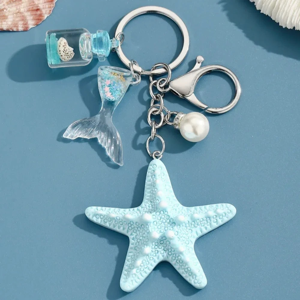 Six pastel resin starfish keychain charms in white, yellow, pink, blue, light pink, and purple, each featuring a mermaid tail, pearl bead, and mini bottle accent on silver chain rings, displayed on a teal background. - 2