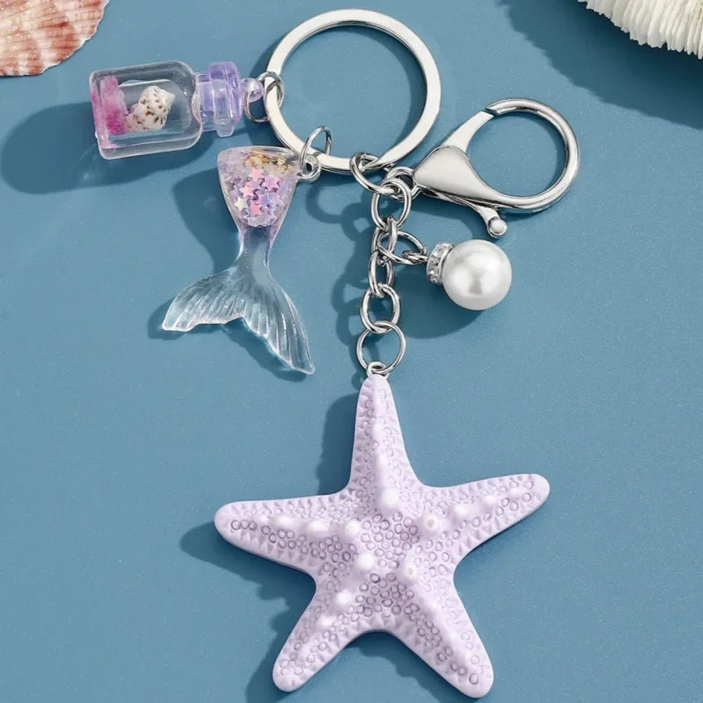 Six pastel resin starfish keychain charms in white, yellow, pink, blue, light pink, and purple, each featuring a mermaid tail, pearl bead, and mini bottle accent on silver chain rings, displayed on a teal background. - 3