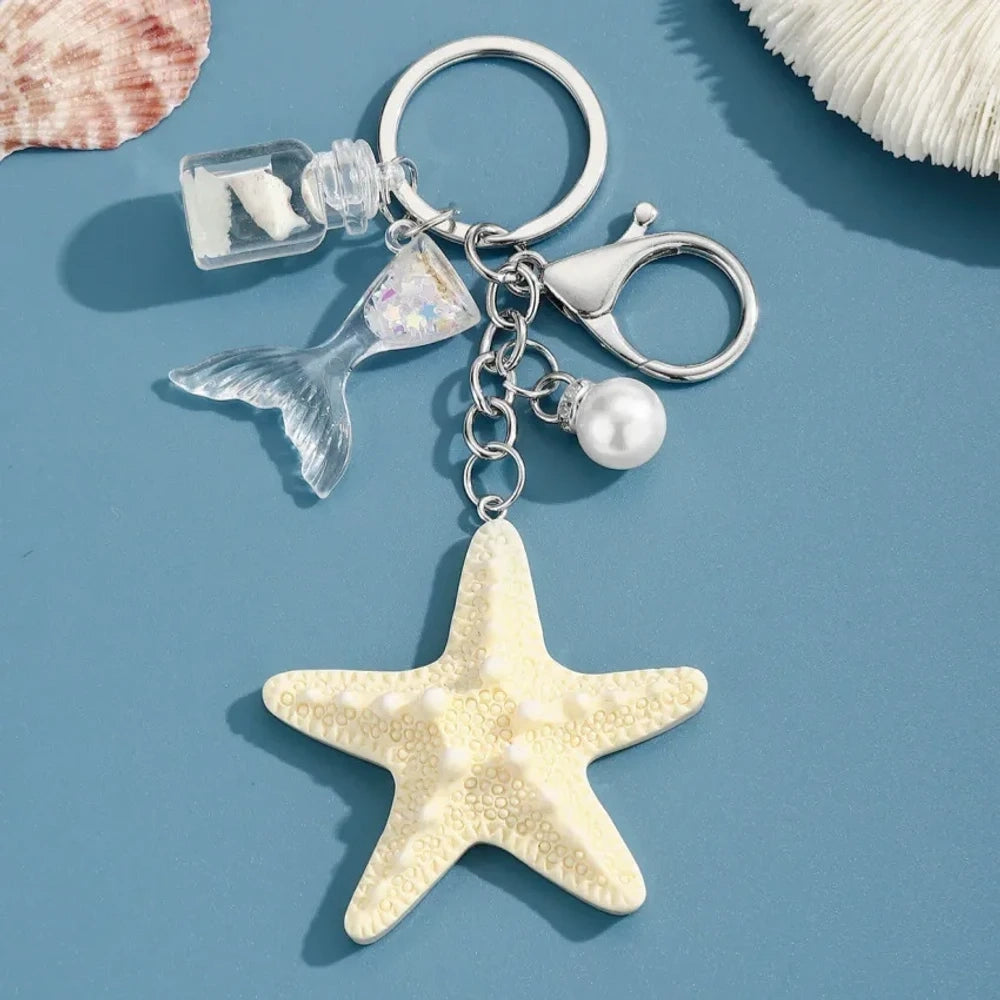 Six pastel resin starfish keychain charms in white, yellow, pink, blue, light pink, and purple, each featuring a mermaid tail, pearl bead, and mini bottle accent on silver chain rings, displayed on a teal background. - 4