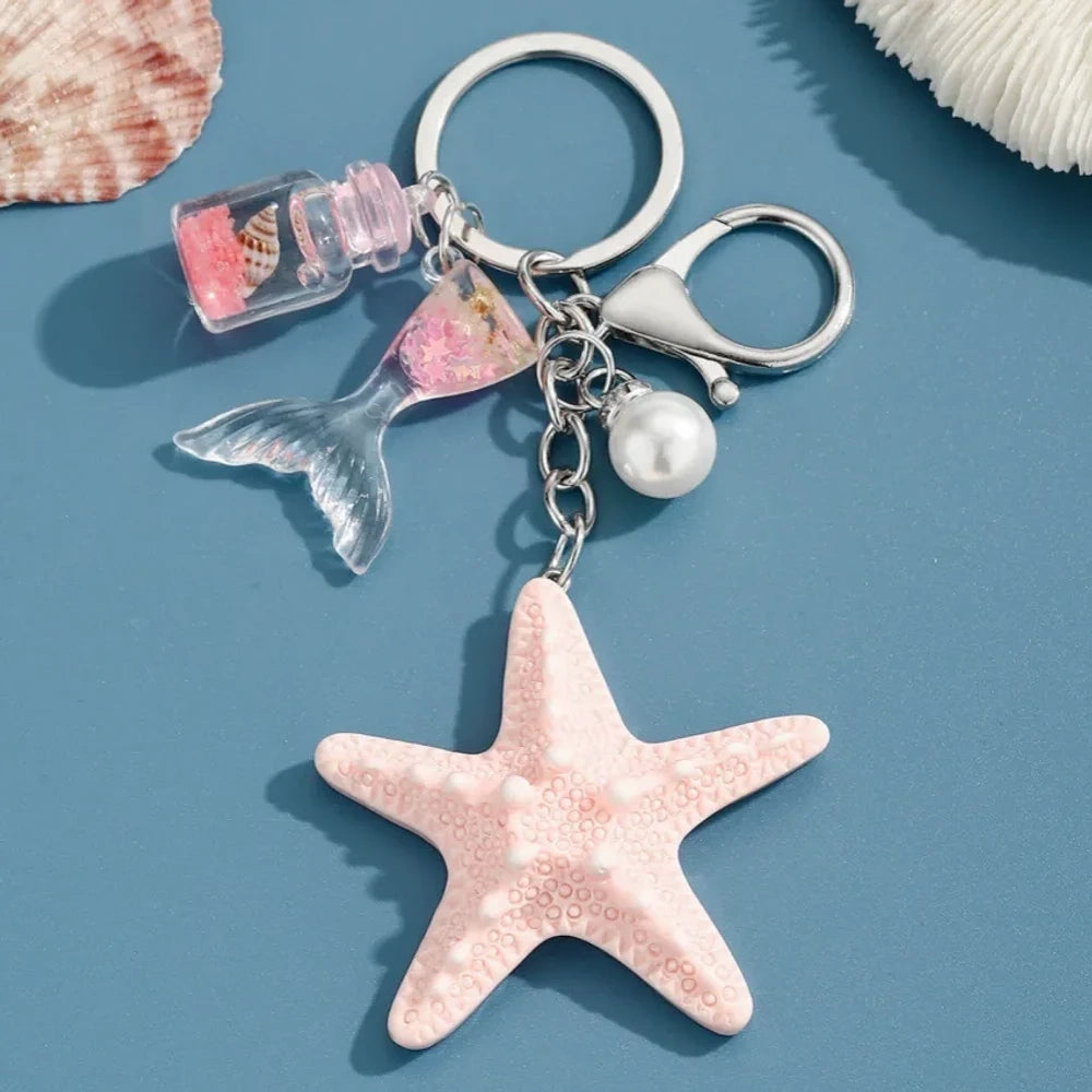 Six pastel resin starfish keychain charms in white, yellow, pink, blue, light pink, and purple, each featuring a mermaid tail, pearl bead, and mini bottle accent on silver chain rings, displayed on a teal background. - 5