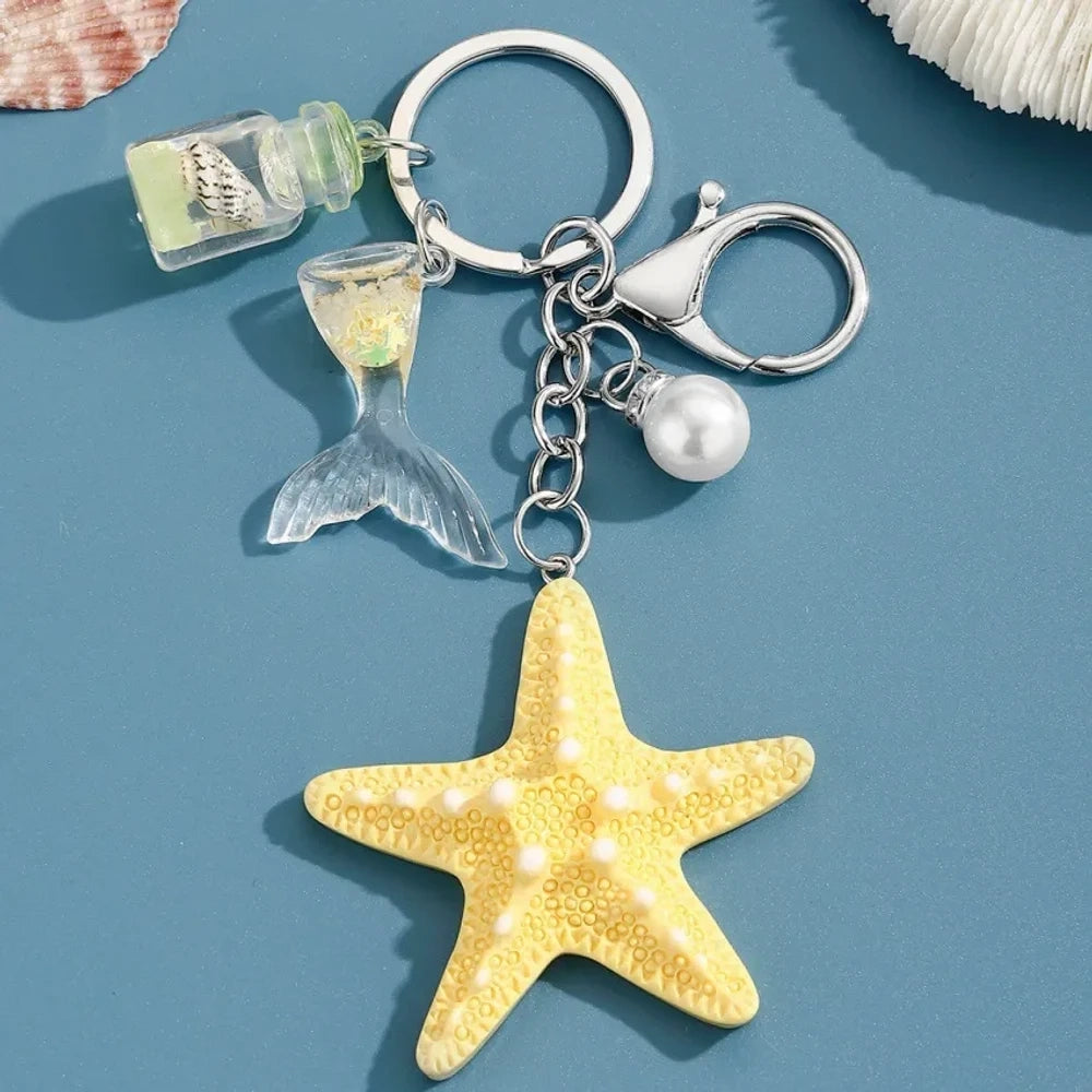 Six pastel resin starfish keychain charms in white, yellow, pink, blue, light pink, and purple, each featuring a mermaid tail, pearl bead, and mini bottle accent on silver chain rings, displayed on a teal background. - 6