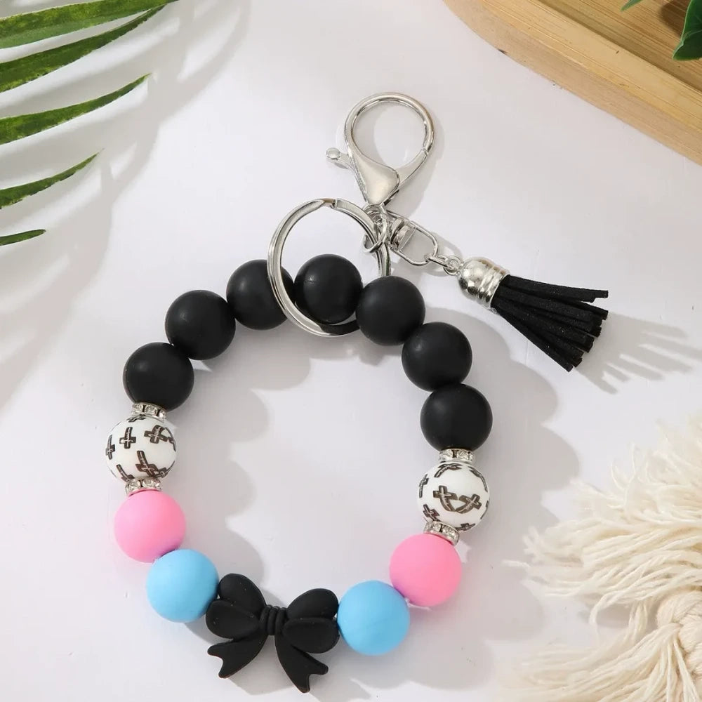 Seven colorful pastel solid silicone bead bracelet keychains in teal, purple, black multicolor, white, pink, and yellow, each featuring bow charms or tassels and silver lobster clasp hardware, arranged on a white surface. - Black