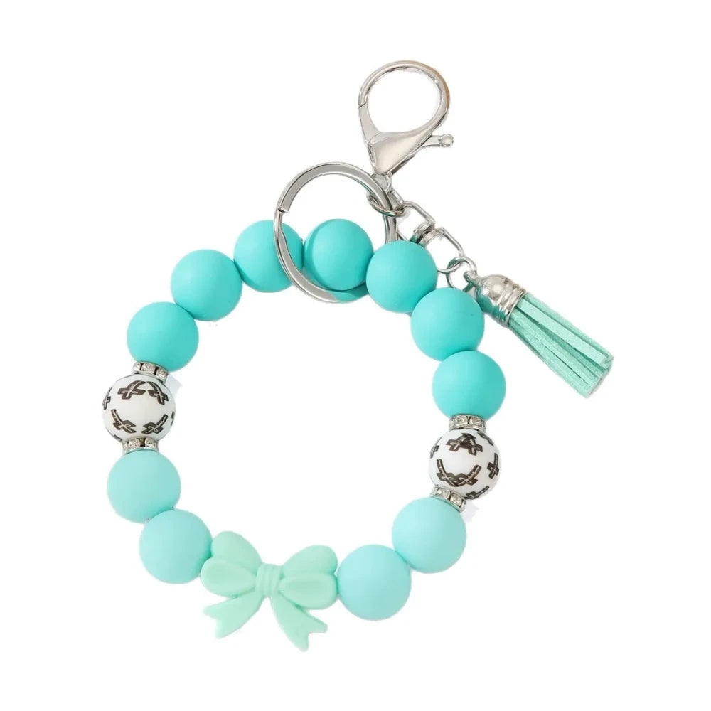 Seven colorful pastel solid silicone bead bracelet keychains in teal, purple, black multicolor, white, pink, and yellow, each featuring bow charms or tassels and silver lobster clasp hardware, arranged on a white surface. - Blue
