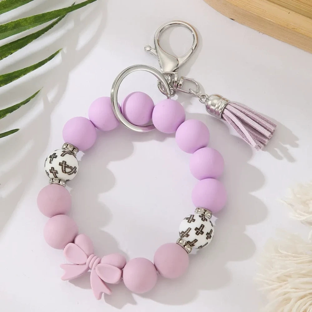 Seven colorful pastel solid silicone bead bracelet keychains in teal, purple, black multicolor, white, pink, and yellow, each featuring bow charms or tassels and silver lobster clasp hardware, arranged on a white surface. - Purple