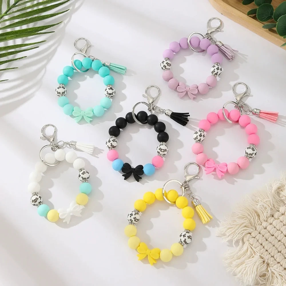 Seven colorful pastel solid silicone bead bracelet keychains in teal, purple, black multicolor, white, pink, and yellow, each featuring bow charms or tassels and silver lobster clasp hardware, arranged on a white surface. - Random Mixed