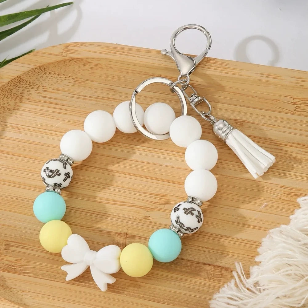 Seven colorful pastel solid silicone bead bracelet keychains in teal, purple, black multicolor, white, pink, and yellow, each featuring bow charms or tassels and silver lobster clasp hardware, arranged on a white surface. - White
