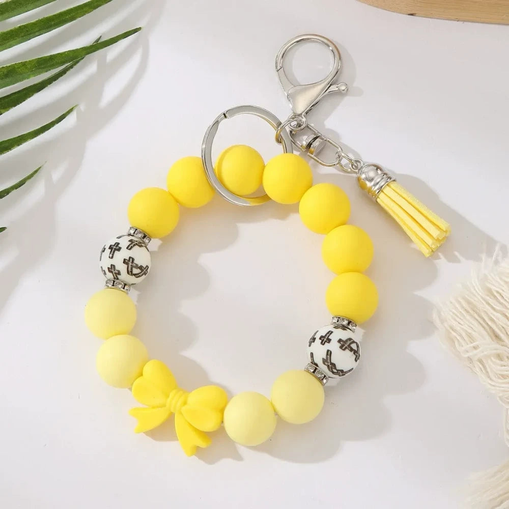 Seven colorful pastel solid silicone bead bracelet keychains in teal, purple, black multicolor, white, pink, and yellow, each featuring bow charms or tassels and silver lobster clasp hardware, arranged on a white surface. - Yellow