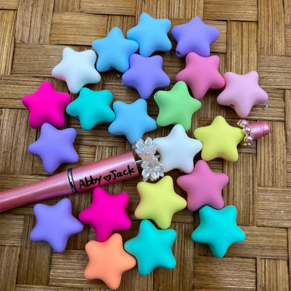 Colorful pastel matte star-shaped acrylic beads in pink, purple, teal, yellow, orange, blue, and green arranged on a wicker surface next to a beadable pen.